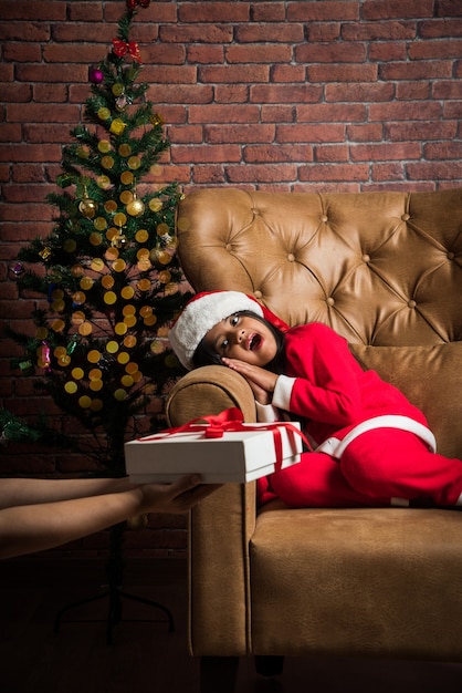 Jolie petite fille asiatique indienne célébrant Noël tout en portant une robe et un chapeau de père Noël, assise sur un canapé avec des cadeaux et un arbre en arrière-plan contre un mur de briques rouges, un éclairage de rêve