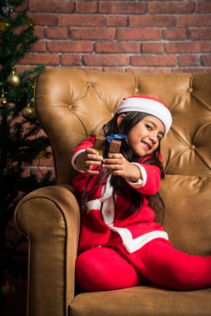 Jolie petite fille asiatique indienne célébrant Noël tout en portant une robe et un chapeau de père Noël, assise sur un canapé avec des cadeaux et un arbre en arrière-plan contre un mur de briques rouges, un éclairage de rêve