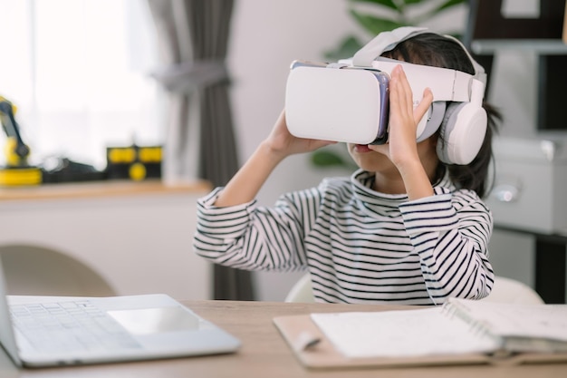 Une jolie petite fille asiatique portant des lunettes VR avec un ordinateur portable placé sur la table dans la classe de technologie STEM Éducation en ligne Érudition