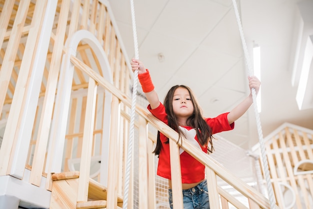 Jolie petite fille asiatique vous regarde en se tenant debout sur un escalier en bois et tenant par des cordes sur l'aire de jeux