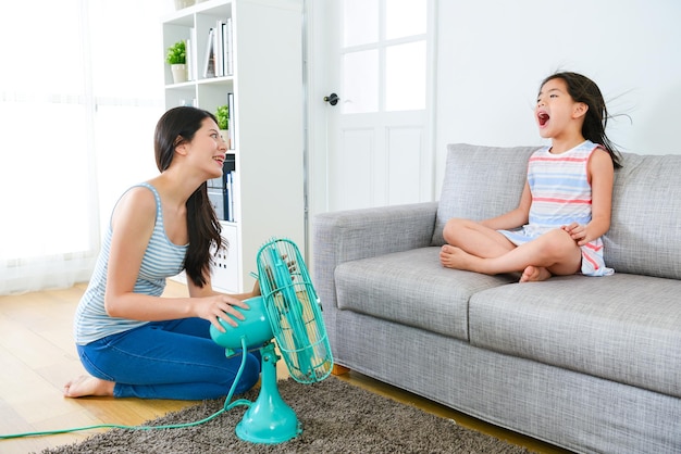 Photo une jolie petite fille assise sur un canapé et une belle femme utilisant un ventilateur électrique lui permettent de profiter d'un vent frais et confortable pendant la saison estivale.