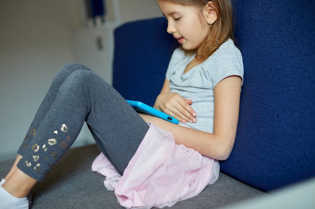 Jolie petite fille assise sur un canapé, enfant accro à la technologie, appréciant de jouer à des jeux en ligne sur une tablette numérique, utilisant des applications, des informations de navigation sur le Web à la maison,