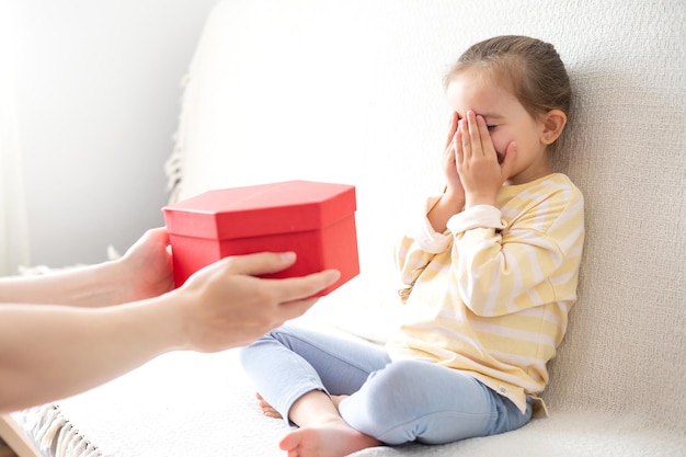 Une jolie petite fille assise sur le canapé et ferme les yeux avec ses mains reçoit un cadeau surprise dans une boîte rouge d'un parent