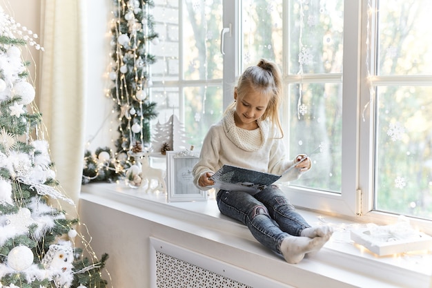 Jolie petite fille assise près de la fenêtre et lisant un livre.