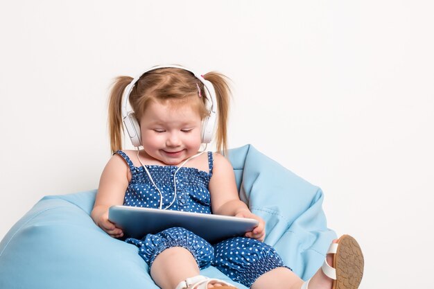 Jolie petite fille au casque écoutant de la musique à l'aide d'une tablette et souriante assise sur un b bleu...