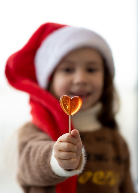 Une jolie petite fille au chapeau du Père Noël tient un bonbon en forme de coeur Nouvel An Noël