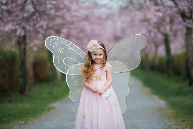 Jolie petite fille aux cheveux bouclés aux cheveux longs dans un beau costume de fée rose avec de grandes ailes dans le jardin de sakura en fleurs Vacances de printemps