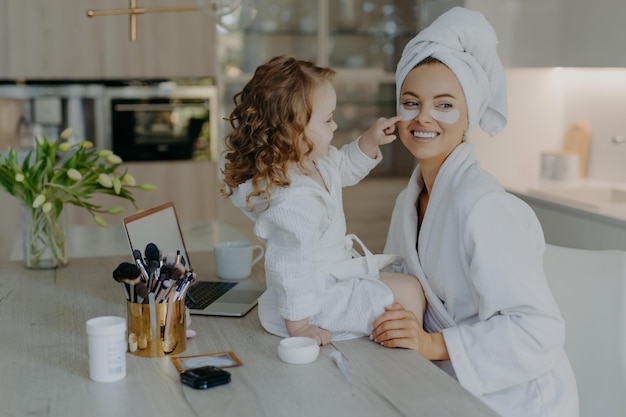 Jolie petite fille aux cheveux bouclés regarde comment la mère fait les procédures cosmétiques applique des patchs de collagène sous les yeux pour le traitement de la peau porte des peignoirs pose près du bureau avec des produits de beauté Soins quotidiens