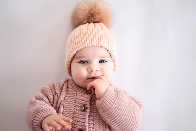 Une jolie petite fille aux yeux bleus dans une combinaison tricotée rose et un chapeau est allongée sur le dos en regardant la caméra sur un fond blanc isolé