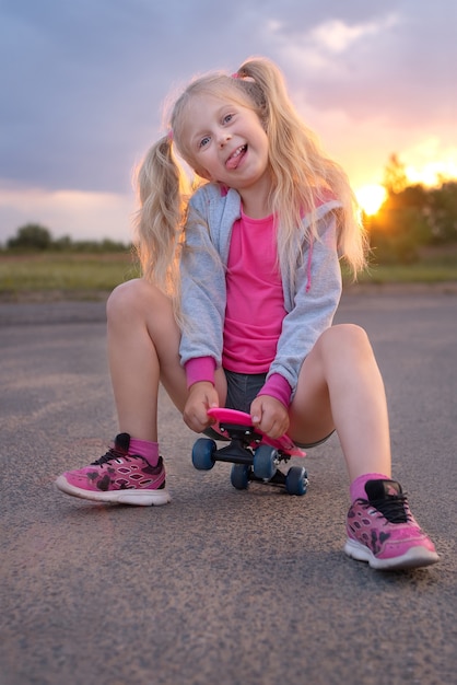 Jolie petite fille blonde assise sur la planche de patinage rose