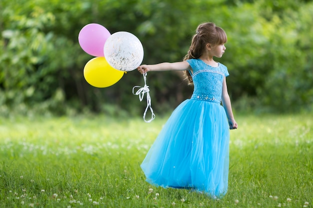 Jolie petite fille blonde aux cheveux longs dans une belle robe de soirée bleue contient des ballons colorés, debout dans un champ fleuri sur floue