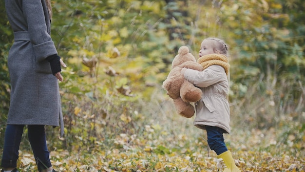 Photo jolie petite fille blonde dans le parc d'automne joue avec un ours en peluche, téléobjectif