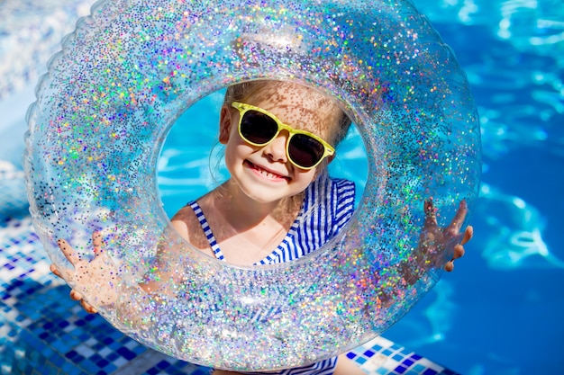 Jolie petite fille blonde à lunettes de soleil rit dans la piscine tenant une bouée de sauvetage