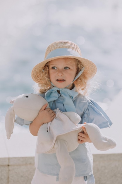 jolie petite fille blonde en robe bleue et chapeau de paille jouant près de la fontaine avec un lapin en peluche, carte