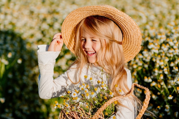 Jolie petite fille blonde vêtue d'une robe en coton et d'un chapeau de paille se promène dans un champ de marguerites les recueille dans le panier