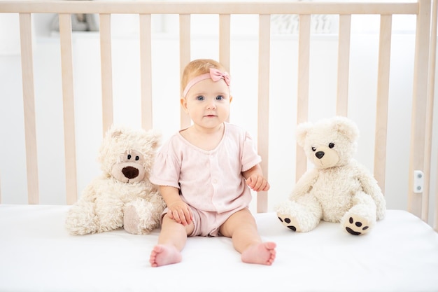 Une jolie petite fille en bonne santé jusqu'à un an dans un body rose en tissu naturel est assise dans un berceau avec des ours en peluche sur une literie blanche dans la chambre en regardant la caméra le bébé est à la maison