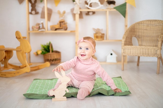 Une jolie petite fille en bonne santé jusqu'à un an dans un costume rose en tissu naturel est assise sur un tapis dans une chambre d'enfants avec des jouets éducatifs en bois regardant la caméra en souriant