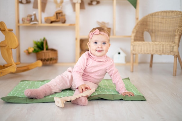 Une jolie petite fille en bonne santé jusqu'à un an dans un costume rose en tissu naturel est assise sur un tapis dans une chambre d'enfants avec des jouets éducatifs en bois regardant la caméra en souriant