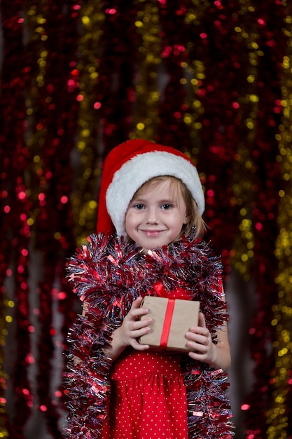 Jolie petite fille en bonnet de Noel et avec des guirlandes autour de son cou tenant le cadeau du nouvel an.