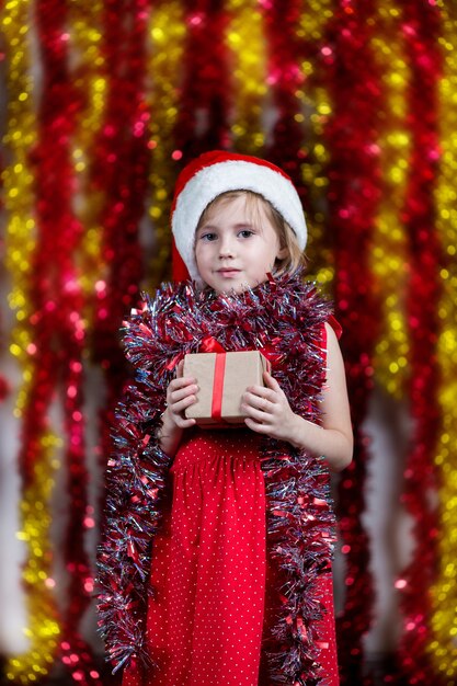 Jolie petite fille en bonnet de Noel et avec des guirlandes autour de son cou tenant le cadeau du nouvel an.