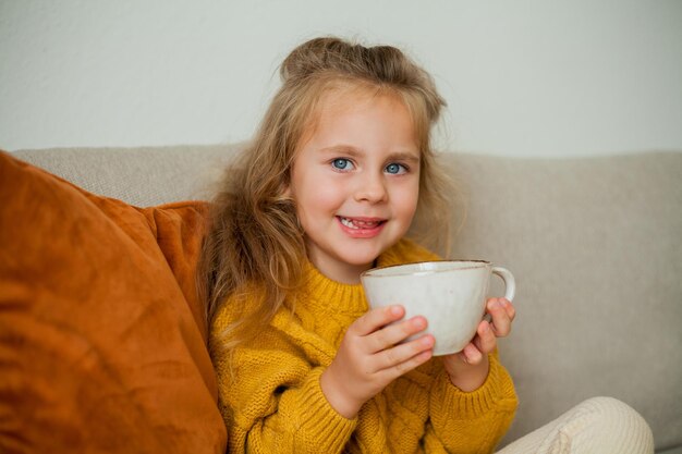 Jolie petite fille bouclée aux yeux bleus de 4 ans buvant du thé dans une maison confortable Boisson chaude d'automne