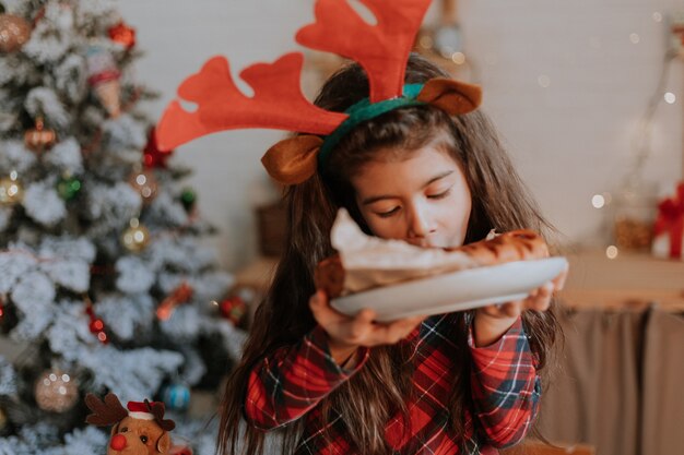 Jolie petite fille brune en pyjama rouge avec des cornes de cerf sur la tête mange un gâteau de Noël