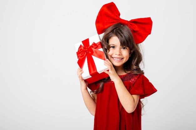 jolie petite fille brune vêtue d'une robe rouge et avec un arc rouge sur la tête tient une boîte-cadeau dans ses mains