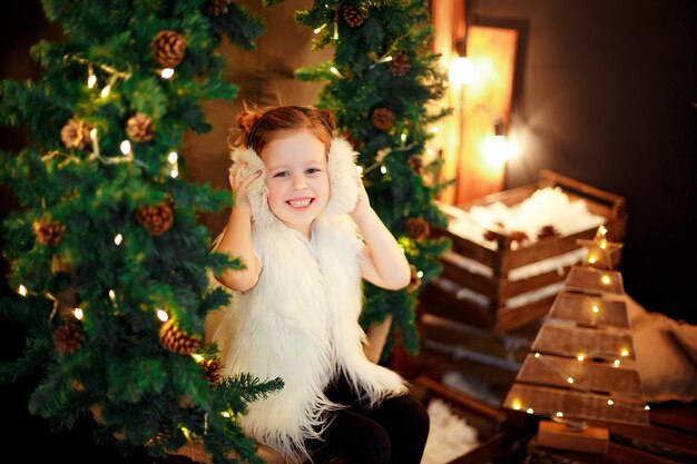 Jolie petite fille en cache-oreilles en fourrure assis près d'un arbre de Noël