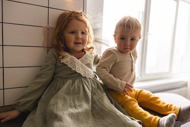 Une jolie petite fille caucasienne et un garçon blond en vêtements décontractés sont assis près de la fenêtre Concept autochtone