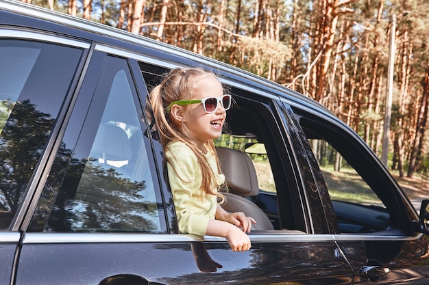 Jolie petite fille caucasienne joyeuse debout à l'intérieur de la voiture en regardant par la fenêtre s'amusant