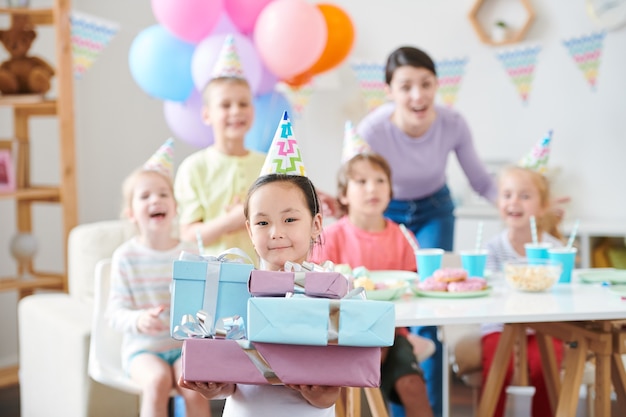 Jolie Petite Fille En Chapeau D'anniversaire Tenant Pile De Cadeaux Amis Extatiques Et Mère S'amusant à La Fête à La Maison