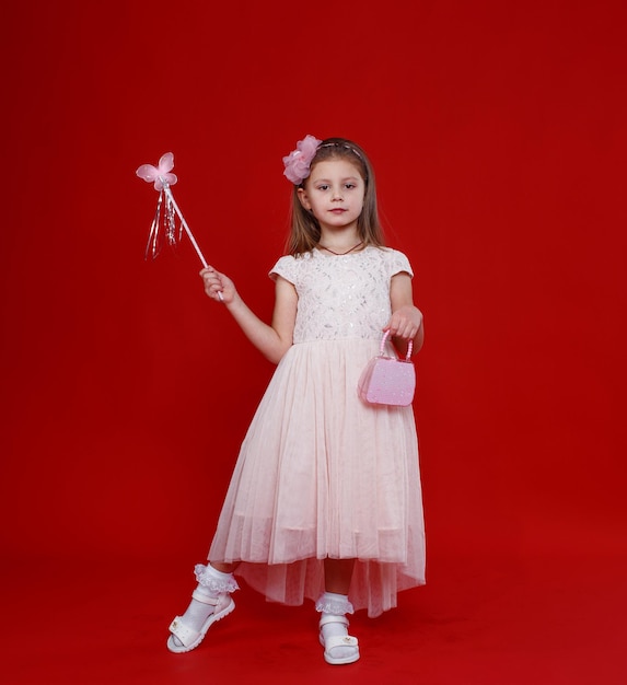 Jolie petite fille en costume de fée rose avec des ailes et une baguette magique sur fond rouge.