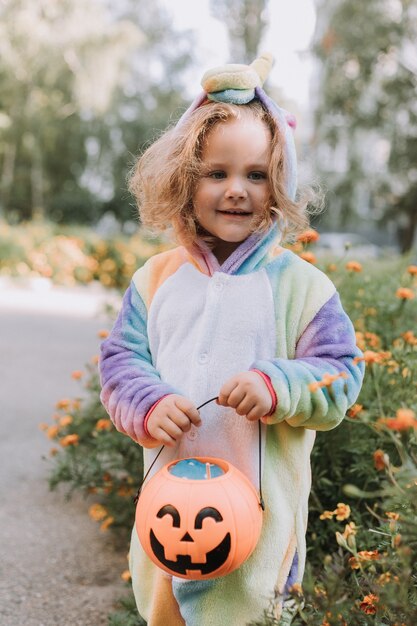 Jolie petite fille en costume de licorne arc-en-ciel pour Halloween va chercher des bonbons dans un panier de citrouilles