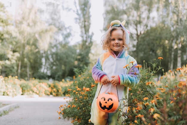 Jolie petite fille en costume de licorne arc-en-ciel pour Halloween va chercher des bonbons dans un panier de citrouilles