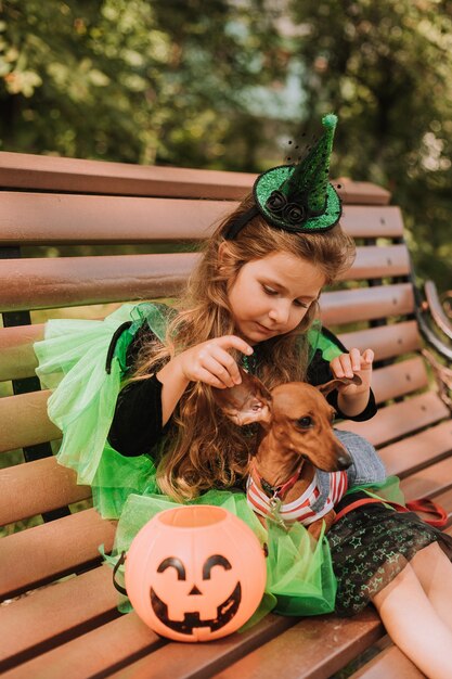 Jolie petite fille en costume de sorcière ou de fée verte d'Halloween est assise sur un banc dans la rue