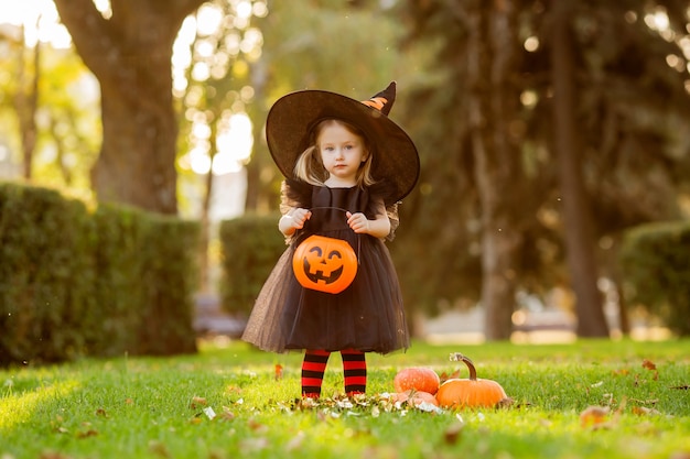 Une jolie petite fille en costume de sorcière se promène dans la rue avec un bonbon en forme de citrouille.