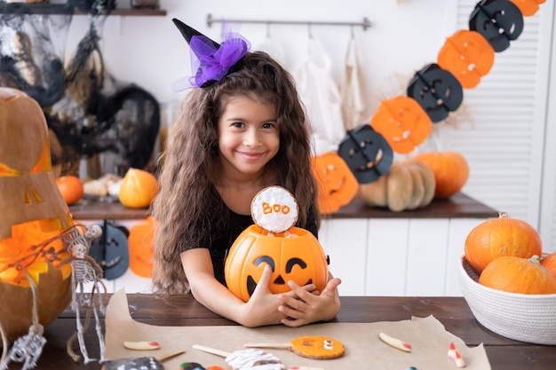 Jolie petite fille en costume de sorcière tenant une citrouille avec des bonbons, s'amusant dans la cuisine, célébrant Halloween.