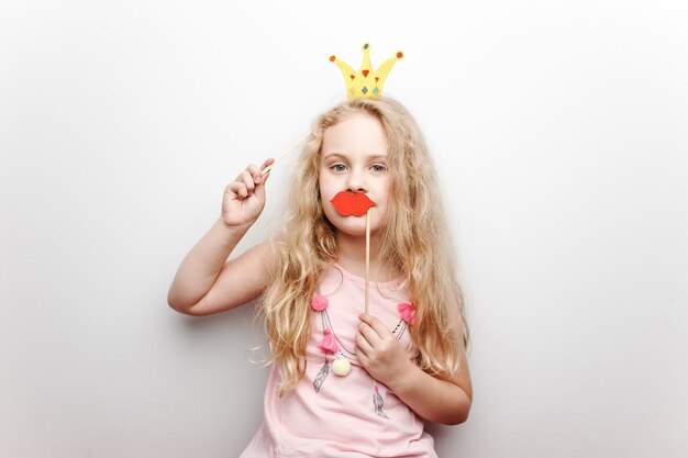 Jolie petite fille avec une couronne de papier et des lèvres rouges est assise sur une chaise rouge à la maison.