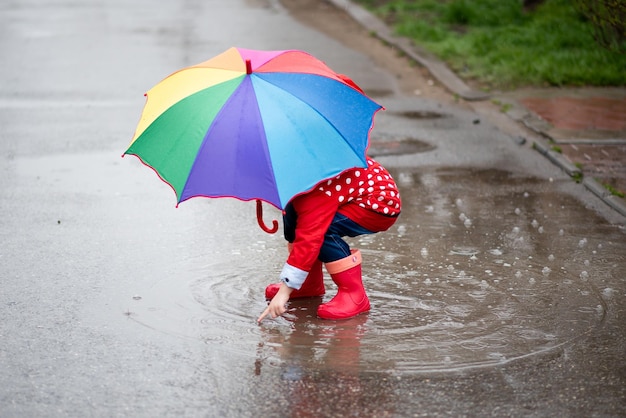 Une jolie petite fille dans une cape rouge, des bottes rouges et un chapeau blanc saute dans les flaques d'eau et s'amuse