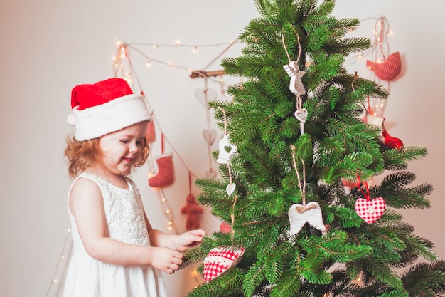 La jolie petite fille dans un chapeau du père Noël décore l'arbre de Noël