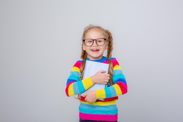 Une jolie petite fille dans un pull multicolore et des lunettes tient un livre sur un fond blanc en souriant