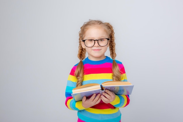 Une jolie petite fille dans un pull multicolore et des lunettes tient un livre sur un fond blanc en souriant