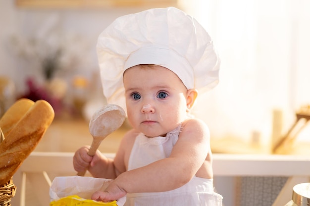 Une jolie petite fille dans une toque de chef et un tablier est assise sur la table dans la cuisine à la maison avec un rouleau à pâtisserie de la farine des miches de pain un petit cuisinier des plats faits maison