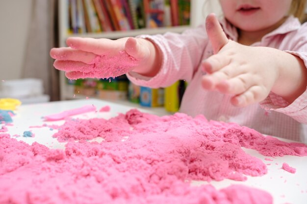 Jolie petite fille enthousiaste joue avec du sable cinétique rose vif qui se trouve sur la table