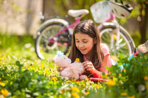 La jolie petite fille est allongée sur une pelouse verte et joue avec un ours en peluche rose tricoté et un pissenlit Son vélo rose en arrière-plan