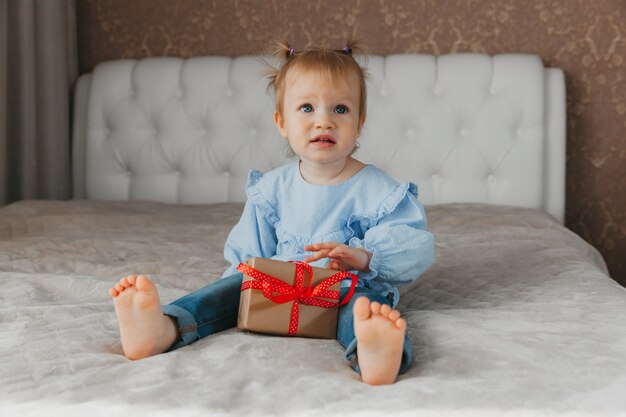 Une jolie petite fille est assise sur le lit avec une boîte-cadeau à la maison, accepte les voeux d'anniversaire.