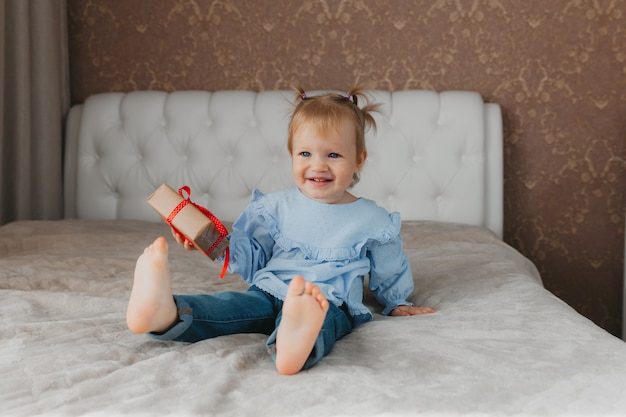 Une jolie petite fille est assise sur le lit avec une boîte-cadeau à la maison, accepte les voeux d'anniversaire.