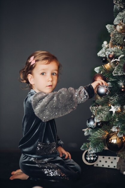 Jolie petite fille est assise sur le sol et décore un arbre de Noël vert à la maison