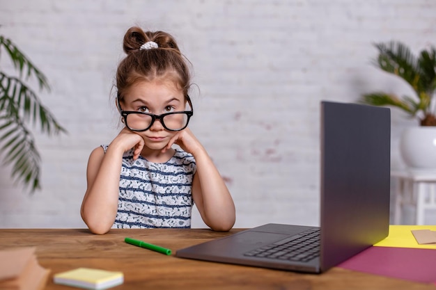 Jolie petite fille est assise à table avec son ordinateur portable et son ordinateur portable, portant des lunettes. Le concept de nouveaux programmes pour enseigner aux enfants