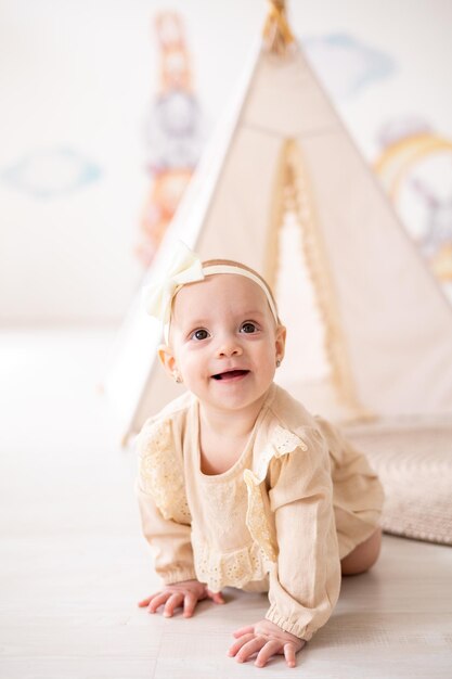 Une jolie petite fille européenne aux yeux bruns dans un body beige en tissu naturel joue dans la chambre des enfants à la maison sur fond de wigwam et de jouets en peluche